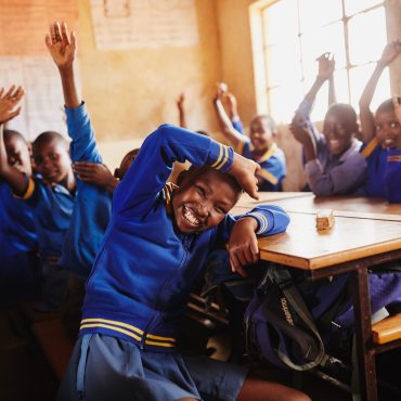 Kinder aus Lesotho sitzen in einem Klassenzimmer erreichtet von World Vision und winken in die Kamera.