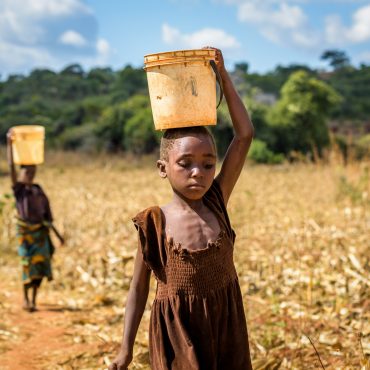 Unterernährtes Mädchen trägt einen schweren Wasserkanister am Kopf und geht durch ein dürres Feld.