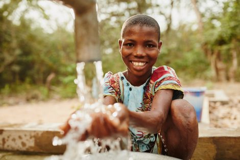Mädchen sitzt lachend vor einer Wasserleitung und lässt sauberes Trinkwasser in ihre Hände fließen.